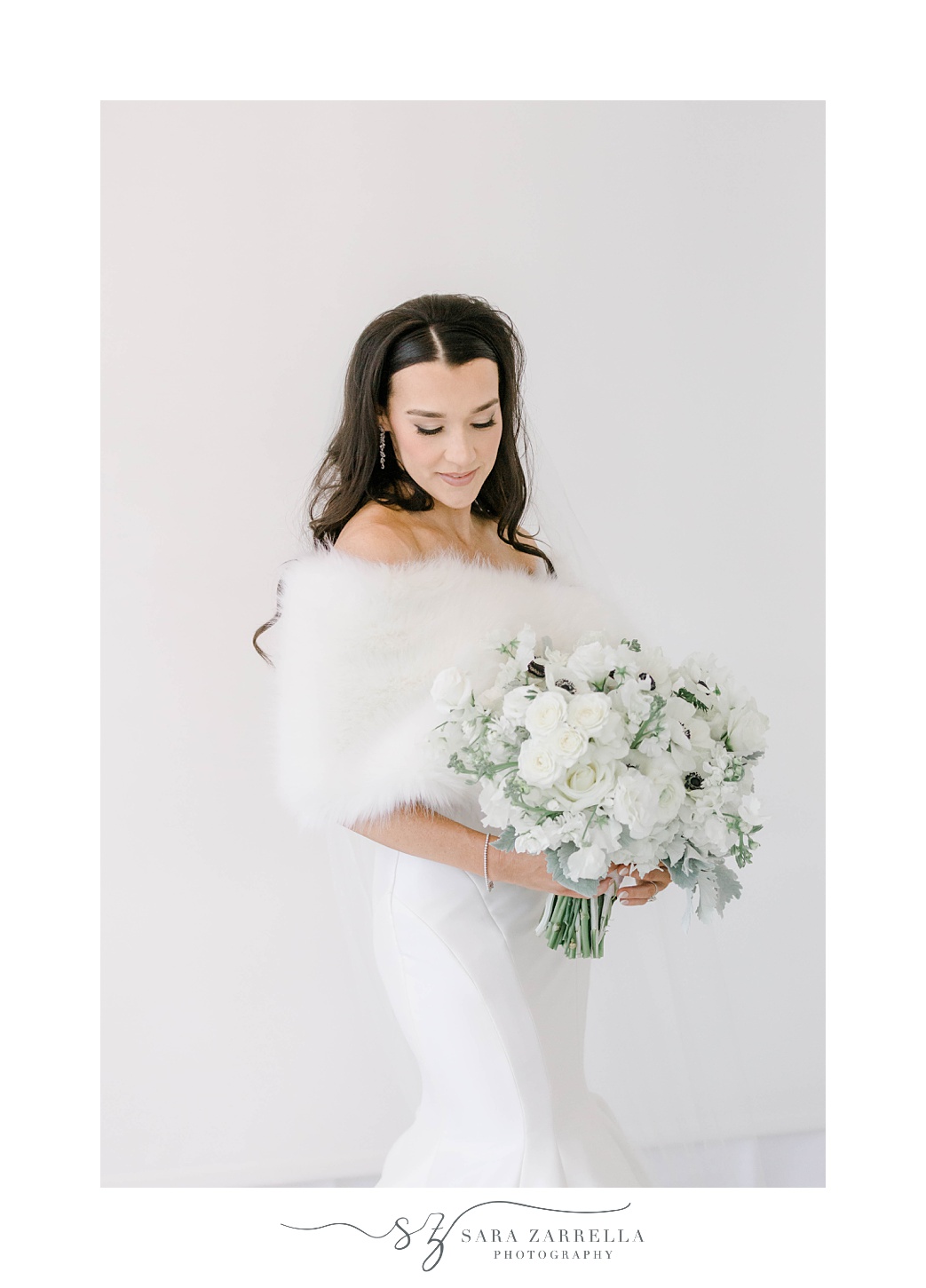 bride in strapless gown and fur looks at bouquet of white anemones