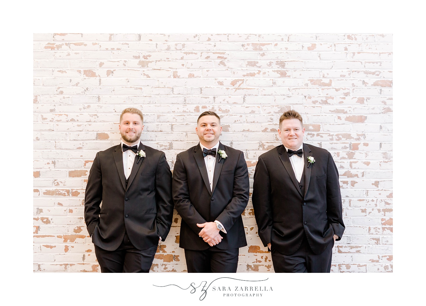 groom and two groomsmen in black suits lean against brick wall in the Crowne Plaza Warwick