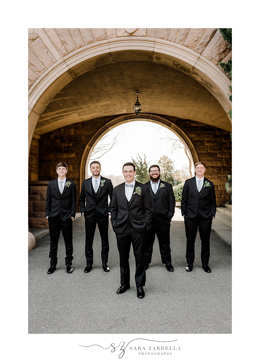 groom and groomsmen in black suits with blue ties pose under archway at OceanCliff Hotel