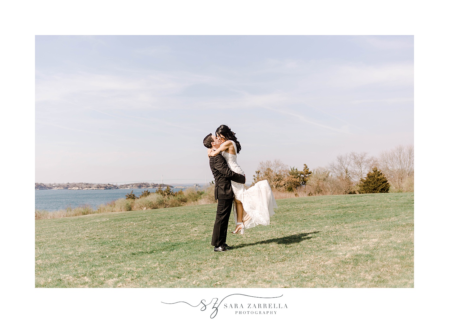 groom lifts bride up and kisses her on hill in Newport RI with water behind them