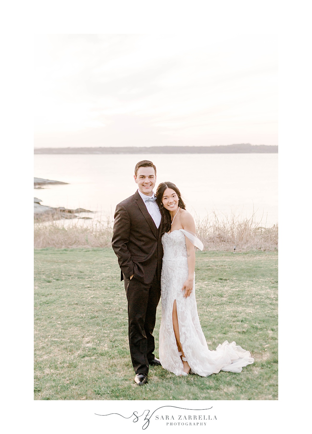 newlyweds stand together hugging on hill in Newport RI with water behind them