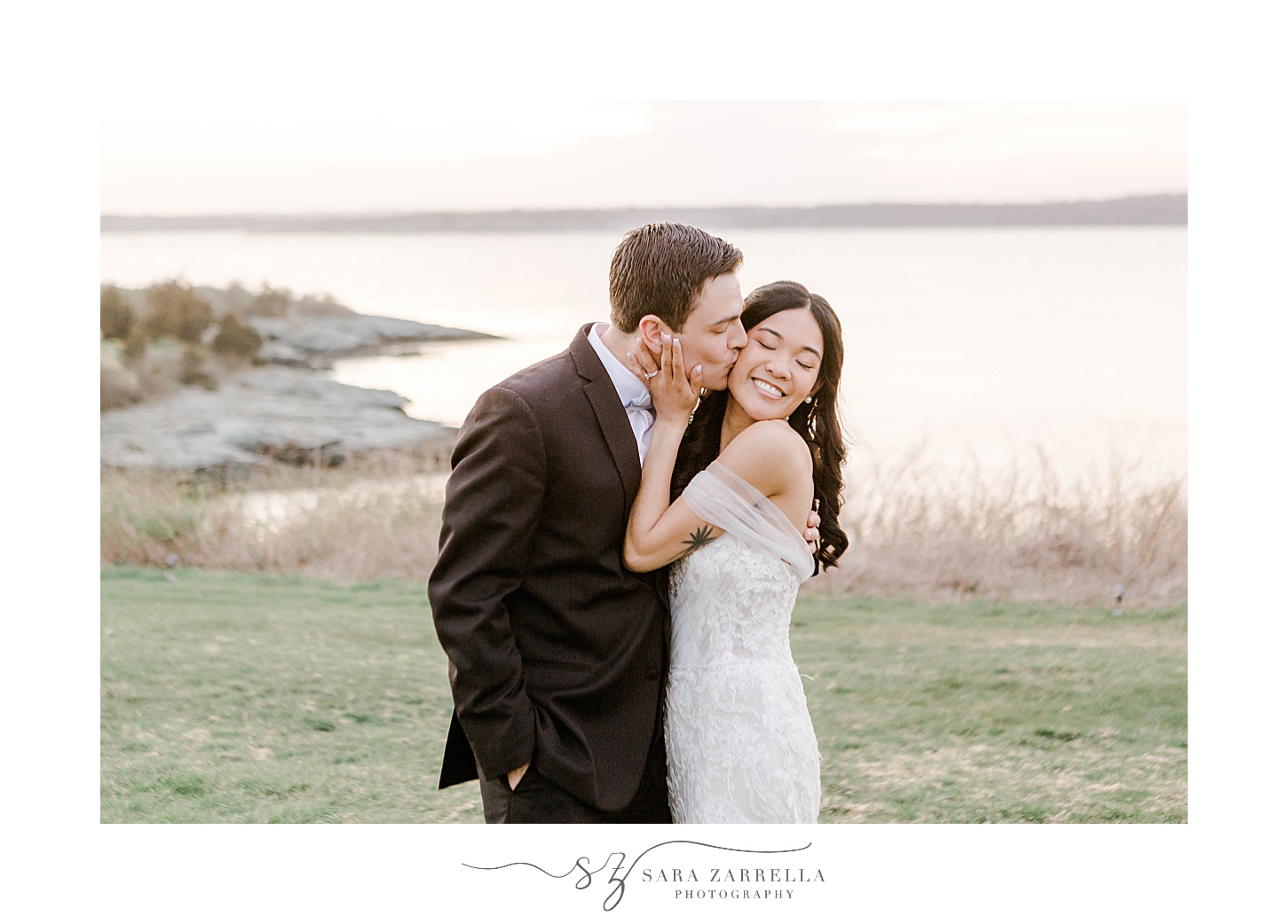 groom kisses bride's cheek on hill in Newport RI with water behind them