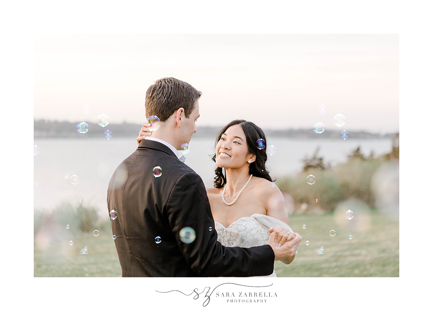 newlyweds dance together at sunset near water while bubbles float around them