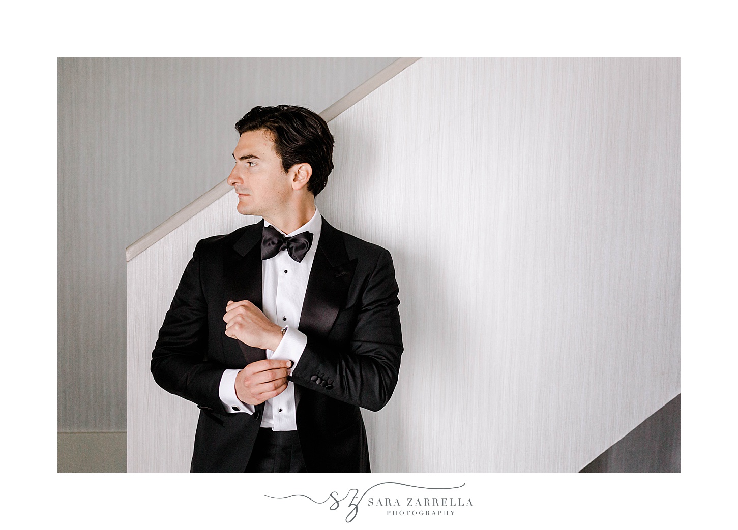 groom adjusts cufflinks of black suit leaning against wall at Belle Mer Island House