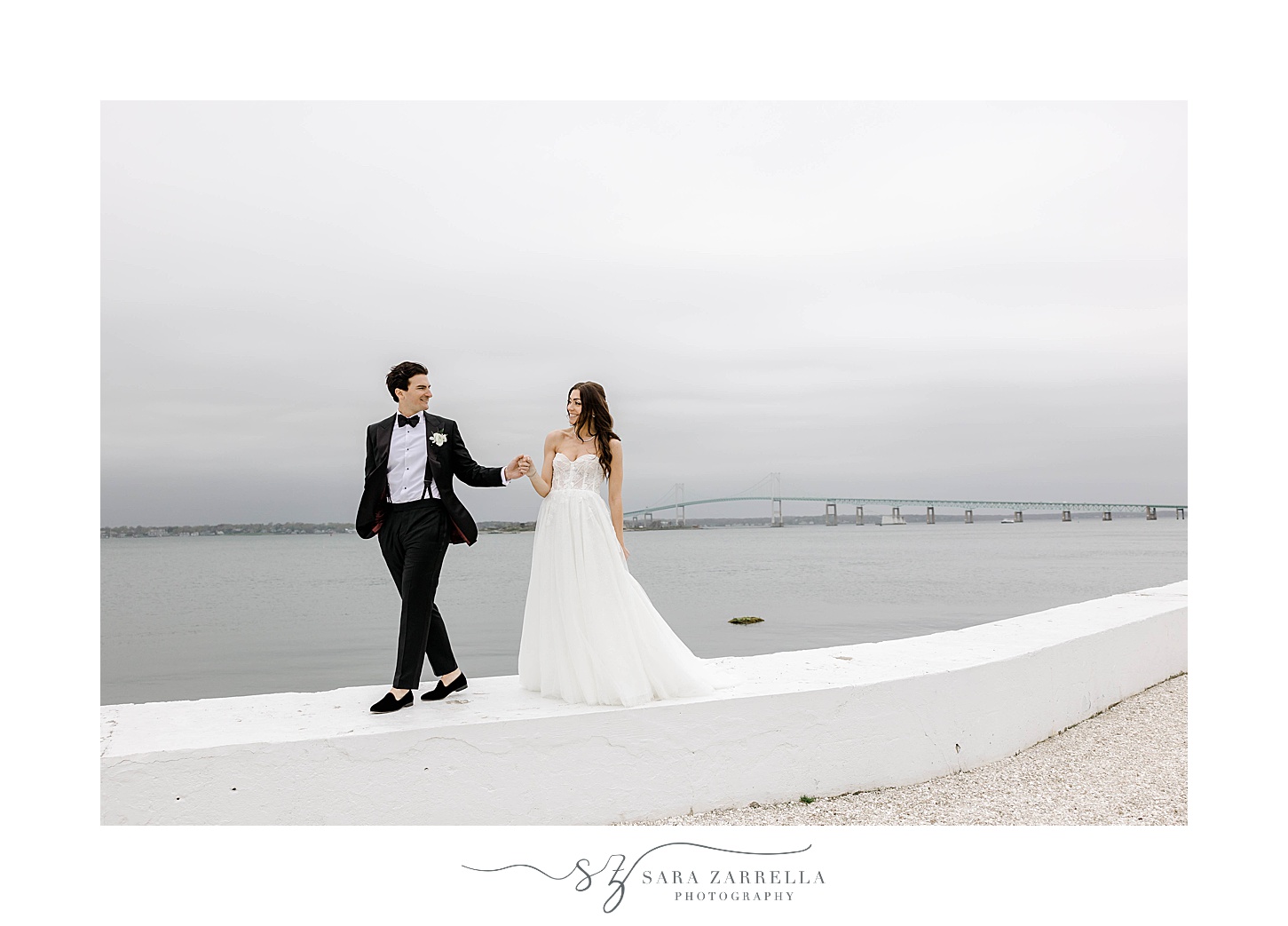 groom walks on wall along Narragansett Bay with bride behind him