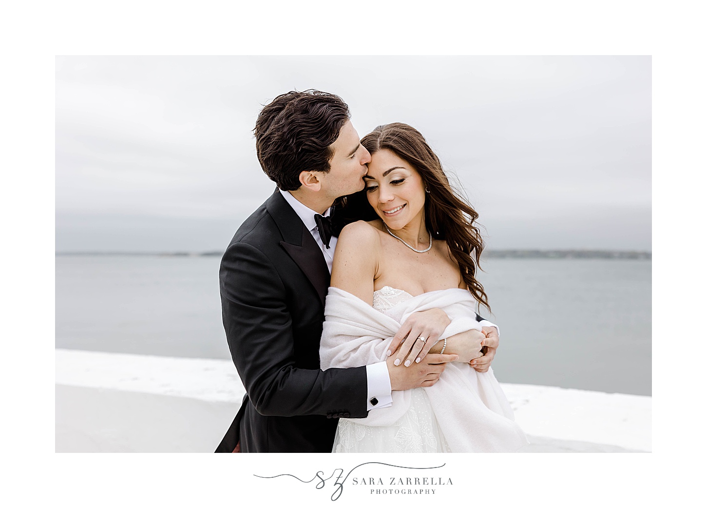 groom hugs bride from behind kissing her forehead by Narragansett Bay 