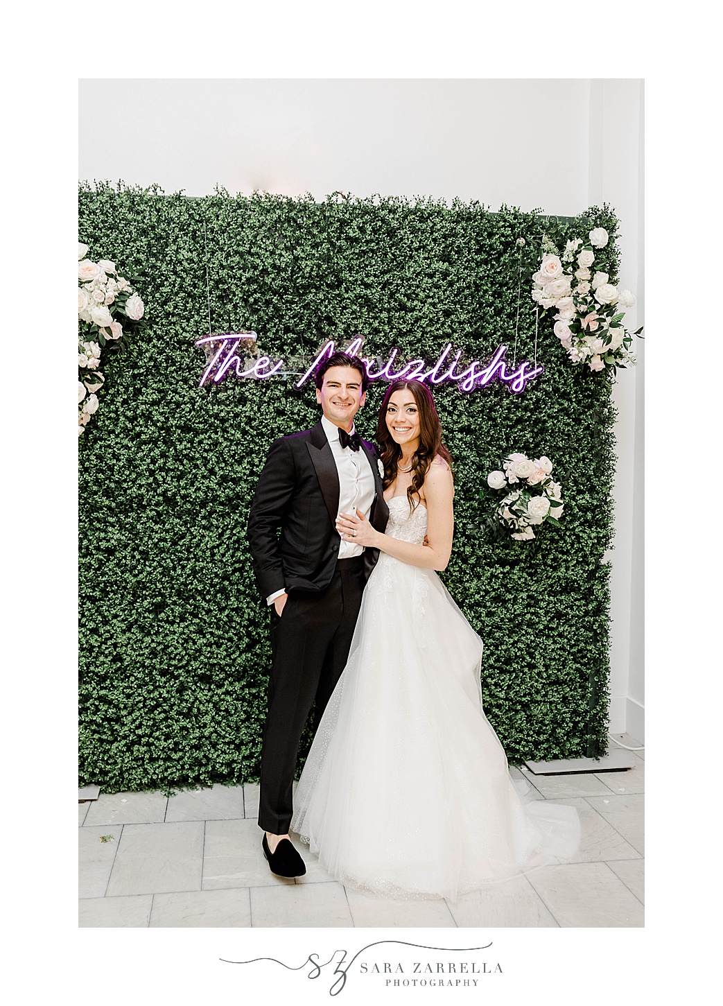 newlyweds pose by greenery wall with custom neon sign