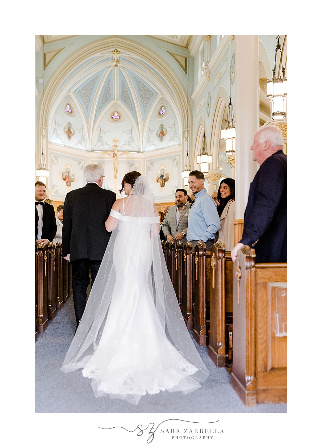 bride walks down aisle with veil trailing behind her inside Our Lady of the Rosary