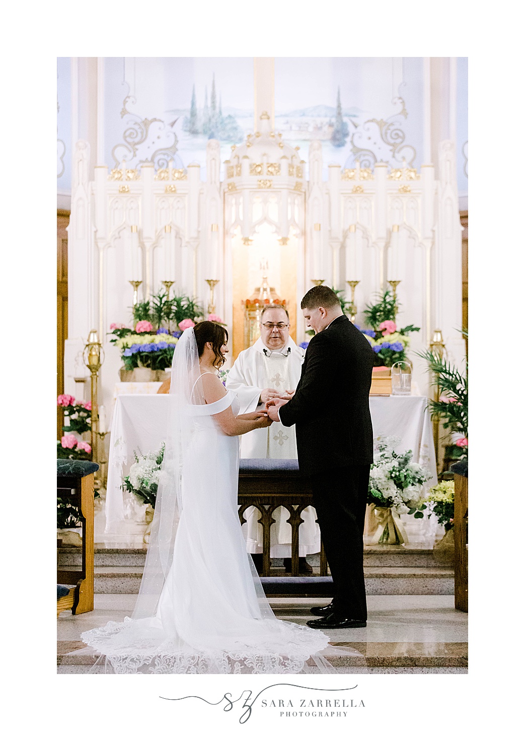 newlyweds hold hands bowing head at alter during wedding ceremony inside Our Lady of the Rosary