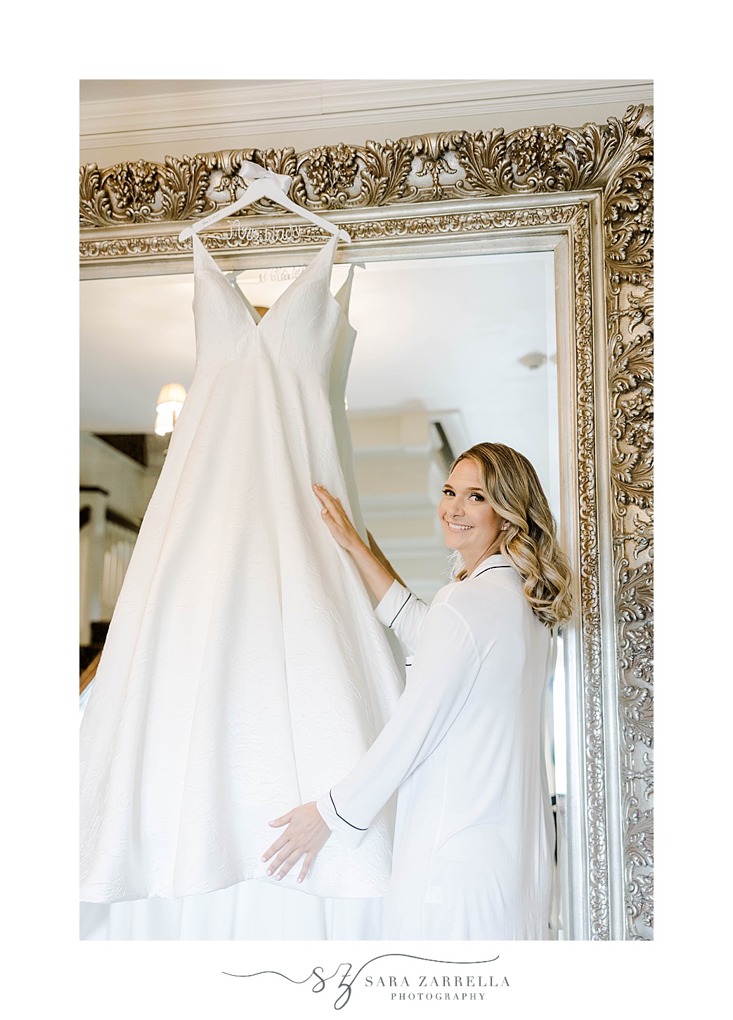 bride looks at wedding dress hanging on gold framed mirror 