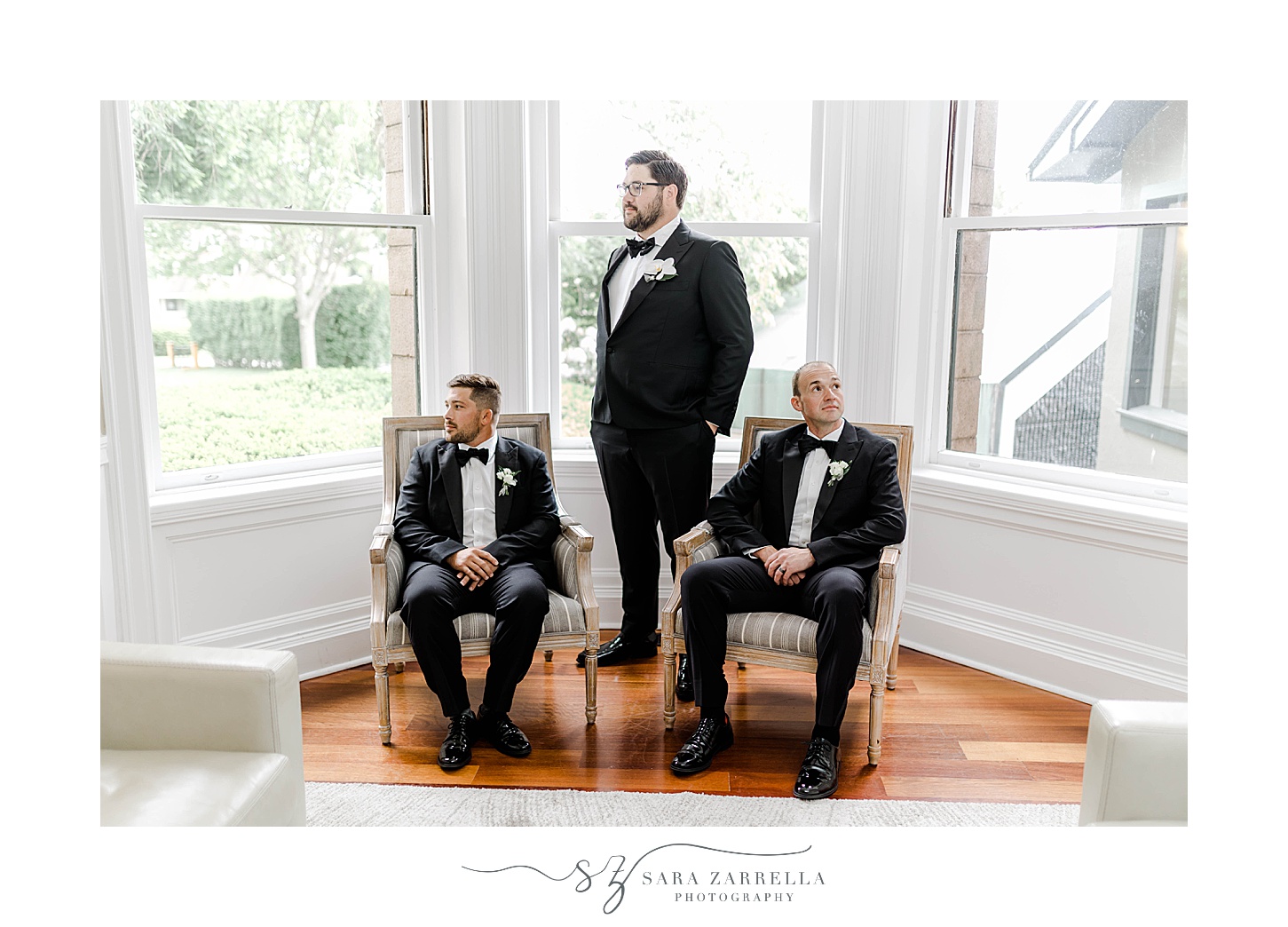 groom stands with two groomsmen in black tuxes in windows of OceanCliff Hotel in Newport RI