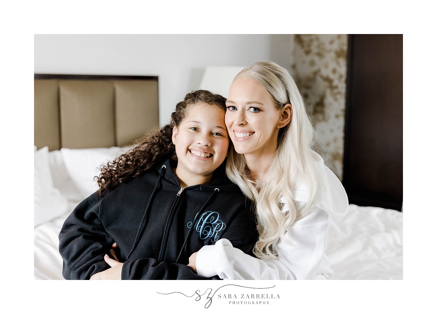 bride sits on bed with flower girl hugging her 