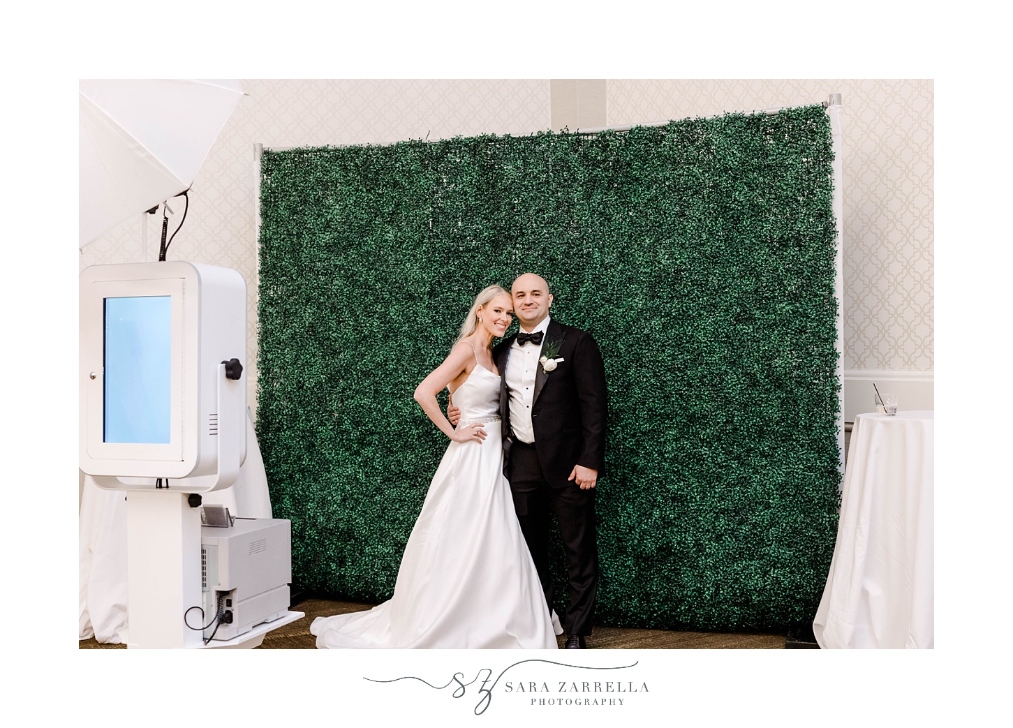 bride and groom pose against greenery wall for Photo Booth 