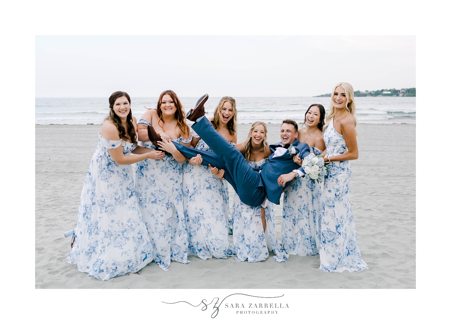 bridesmaids in blue and white dresses hold groom in blue suit on Newport Beach