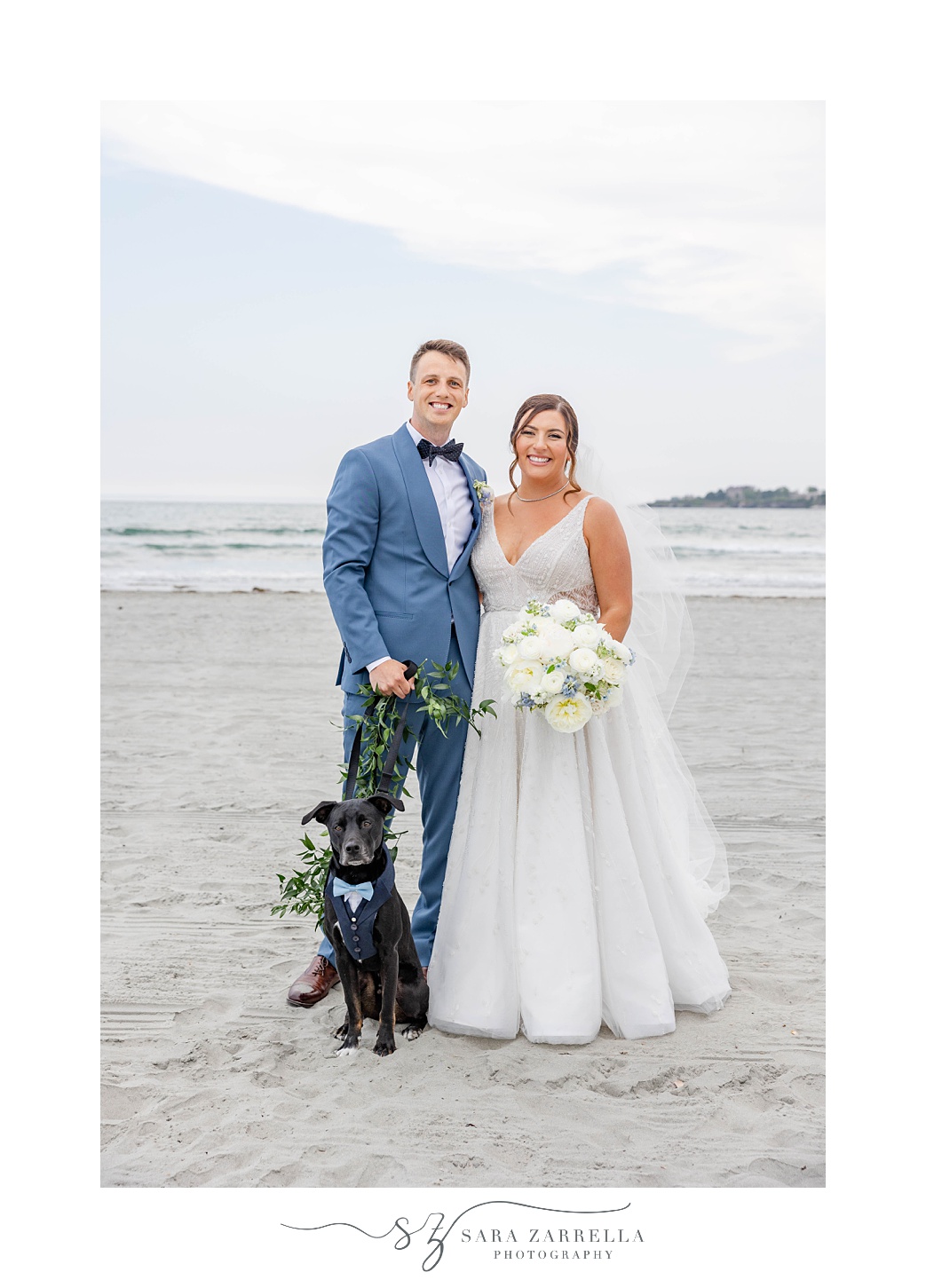 bride and groom stand on beach with black lab puppy