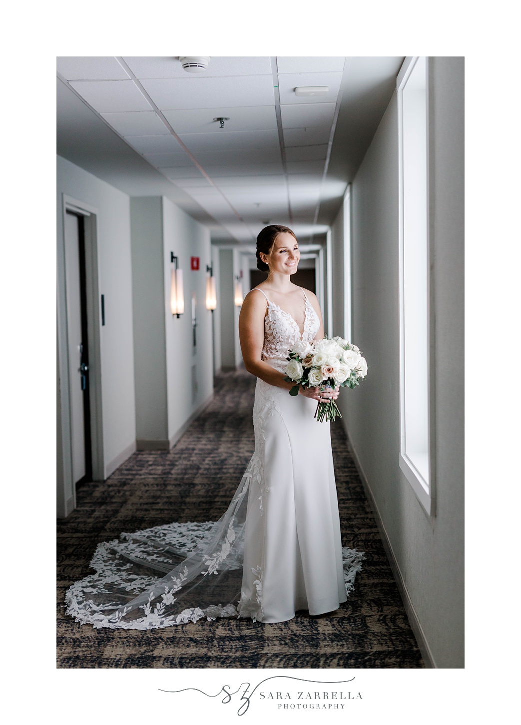bride stands in hallway at OceanCliff Hotel looking out window with bouquet of flowers 