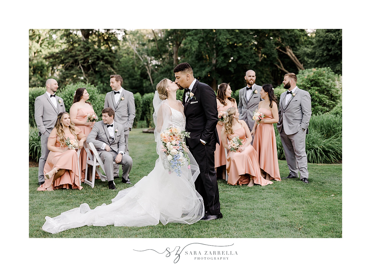 newlyweds kiss while groomsmen in grey suits and bridesmaids in pink gowns stand on lawn 