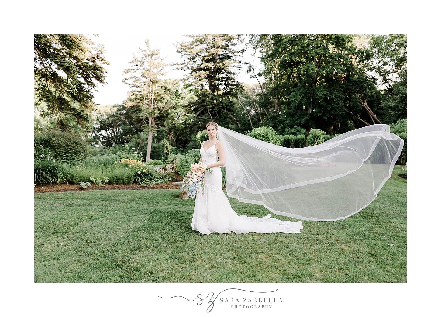 bride smiles holding bouquet standing on lawn with veil behind her