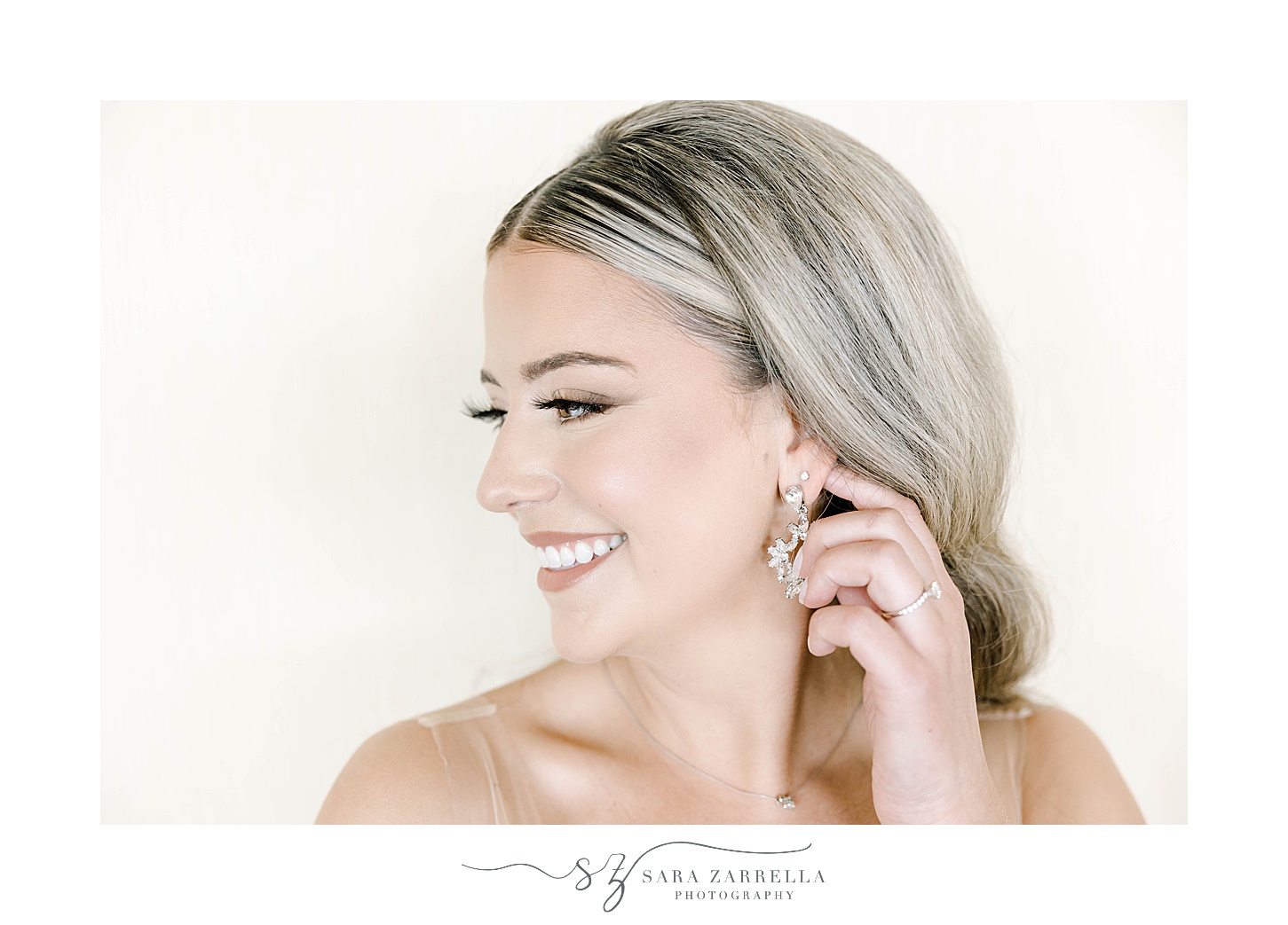 bride shows off diamond earrings in front of ivory wall 