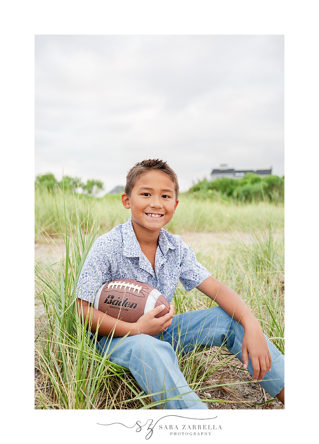 boy sits in tall grass holding football on Rhode Island beach
