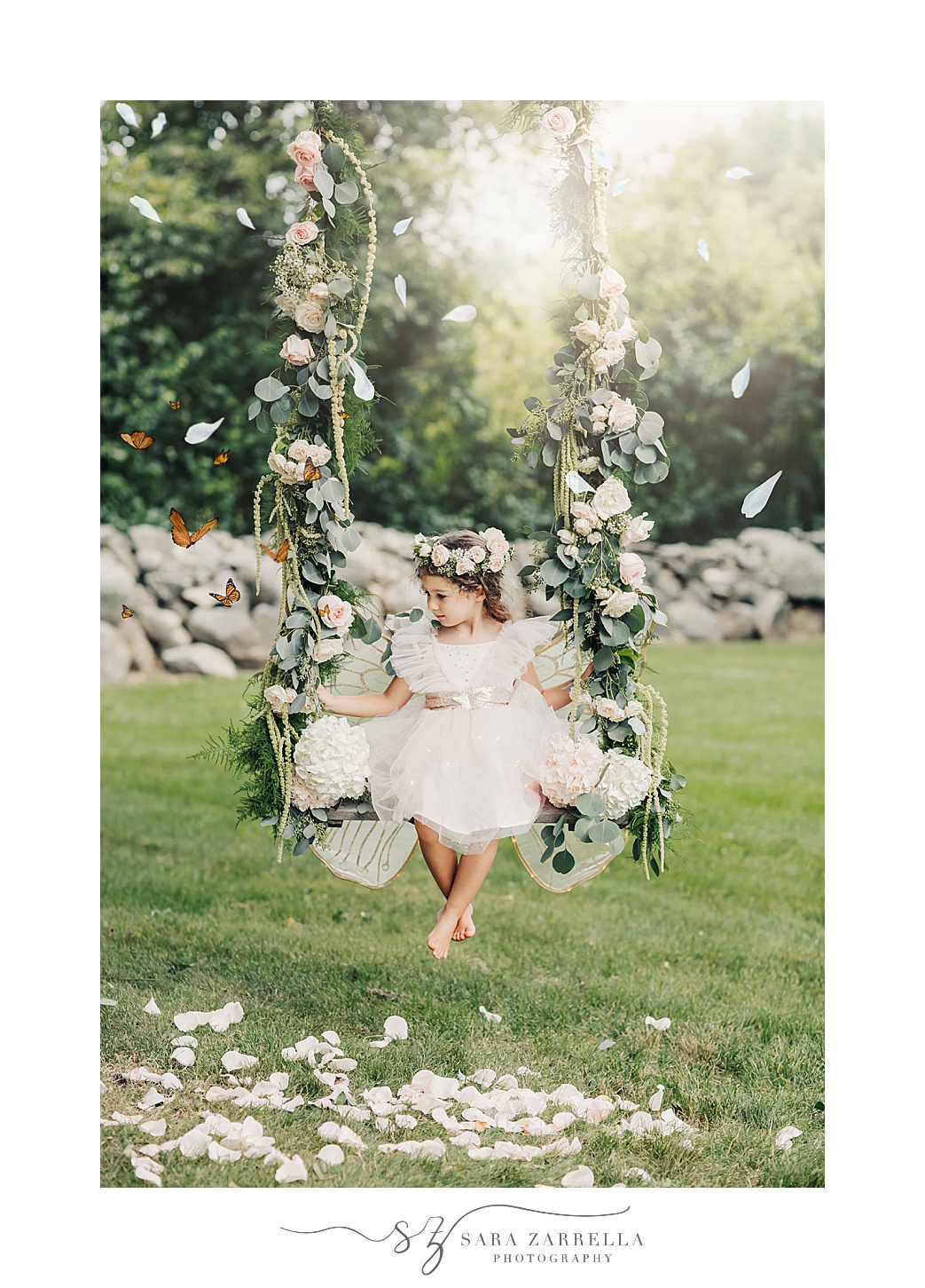 girl in fairy wings and flower crown swings on wooden swing during summer mini session on farm in Rhode Island