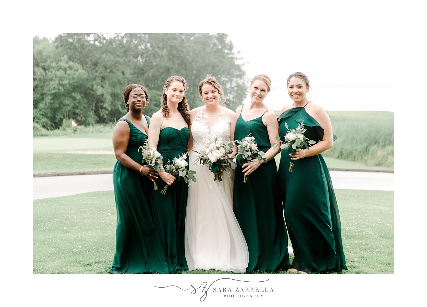 bride poses with bridesmaids in emerald gowns on hill at Harbor Lights