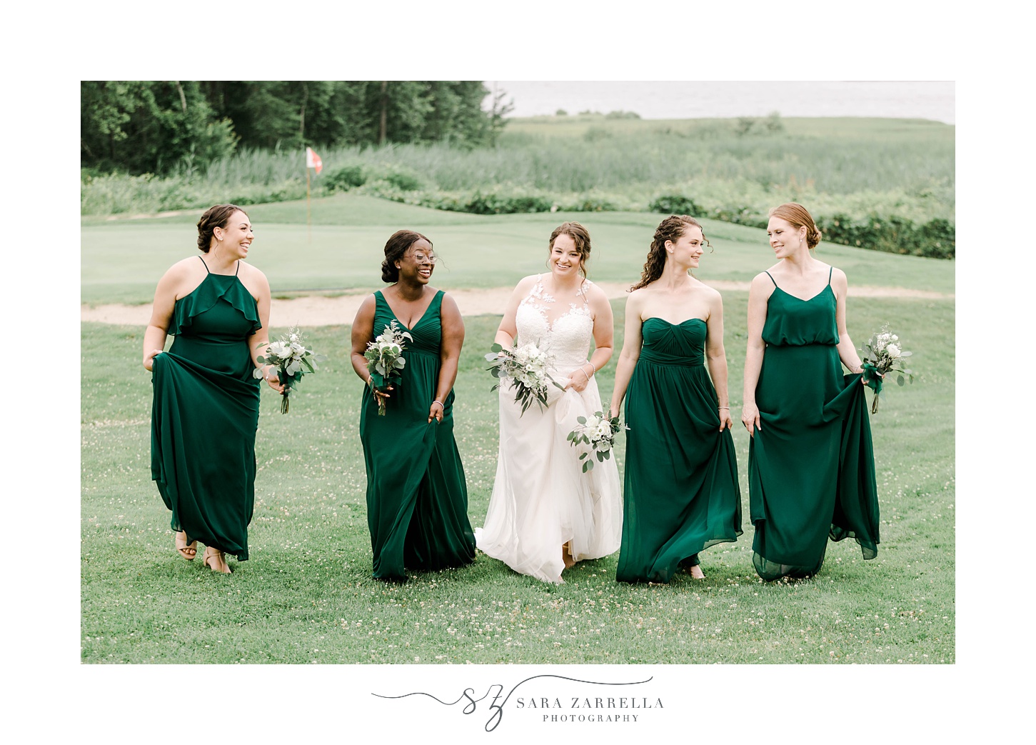 bride and bridesmaids in dark green gowns walk on hill at Harbor Lights