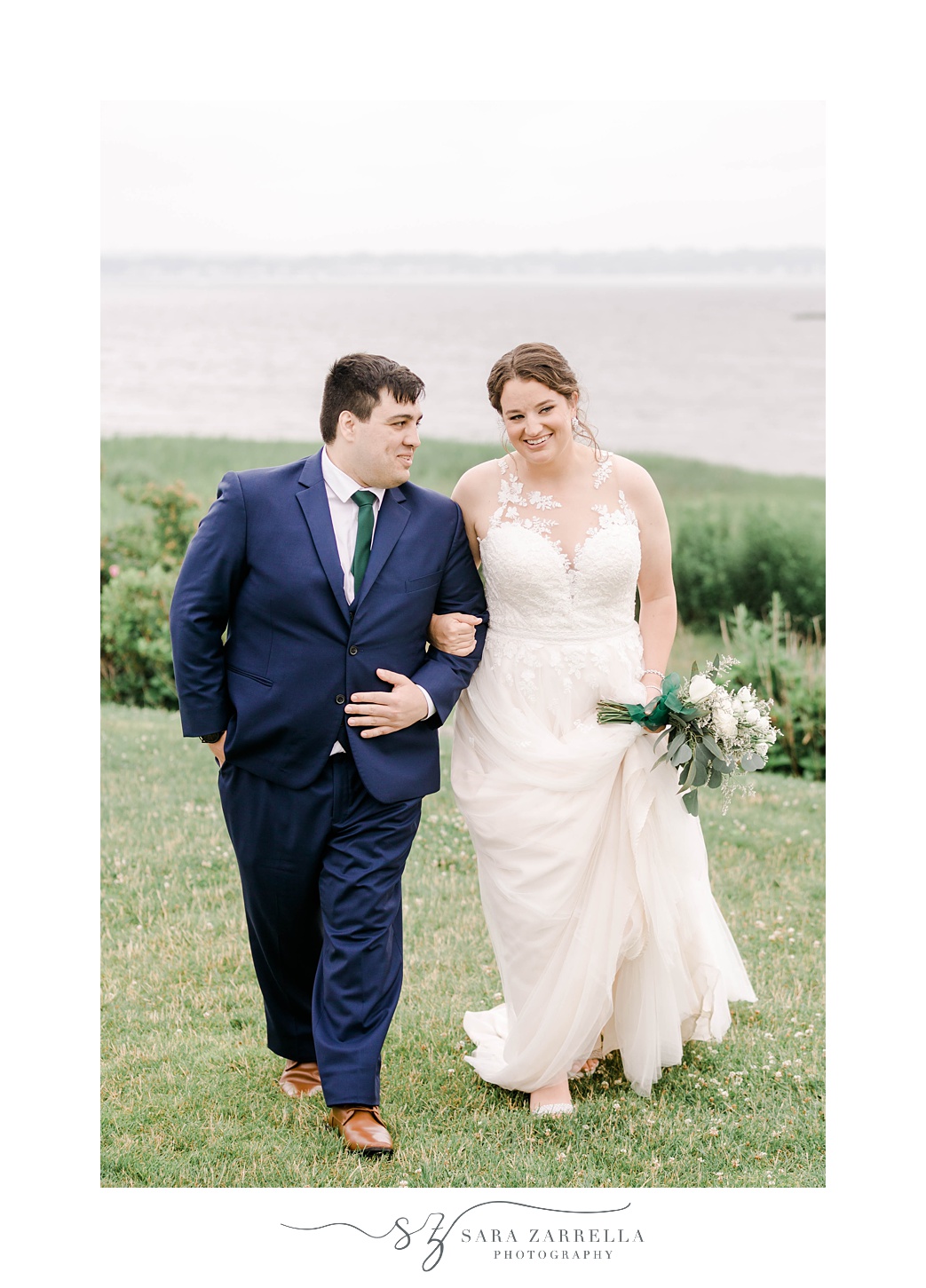 bride walks with groom, placing her arm in his with Narragansett Bay behind them