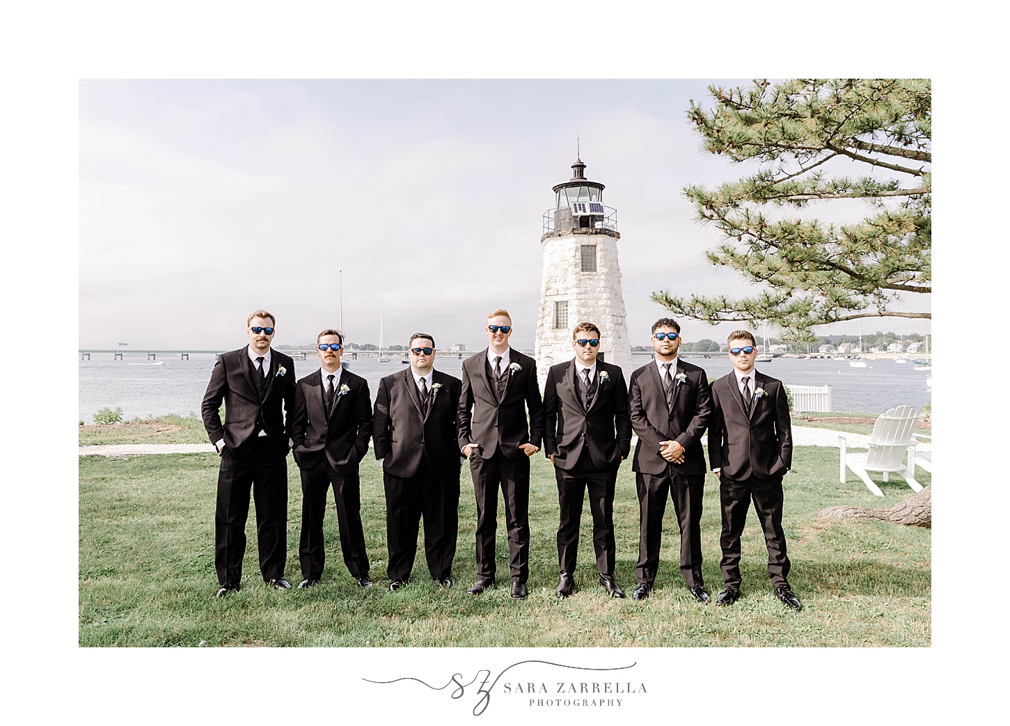 groom poses with groomsmen in black suits in front of Newport Lighthouse 