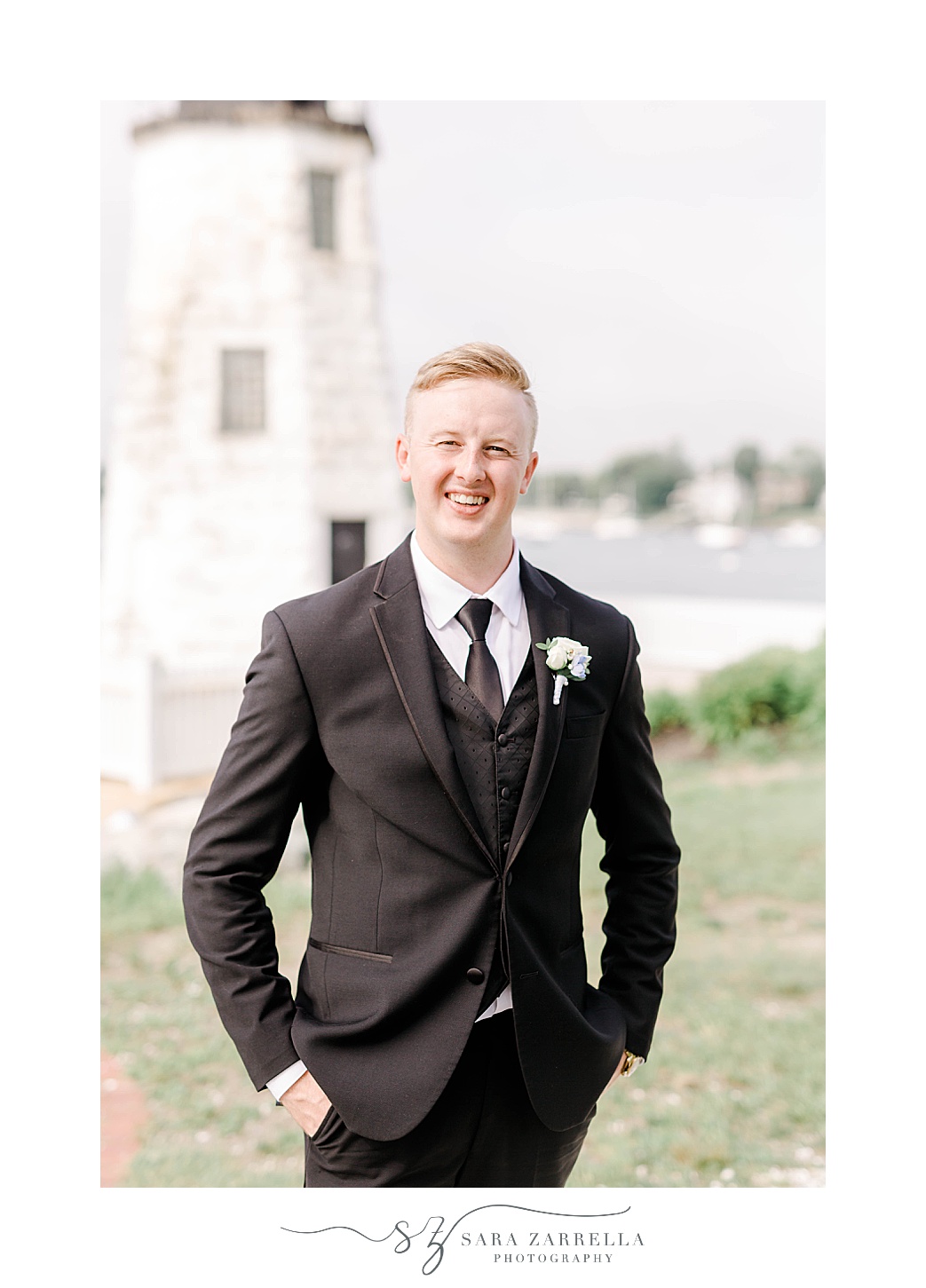 groom stands with hands in pocket of black suit in front of Newport Lighthouse 