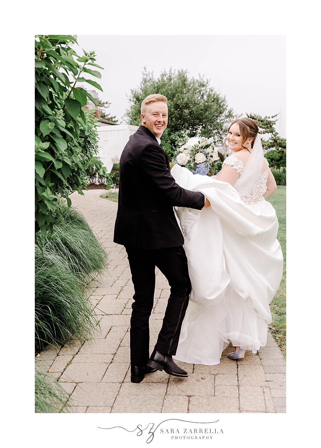 groom helps bride carry wedding dress walking on brick walkway 