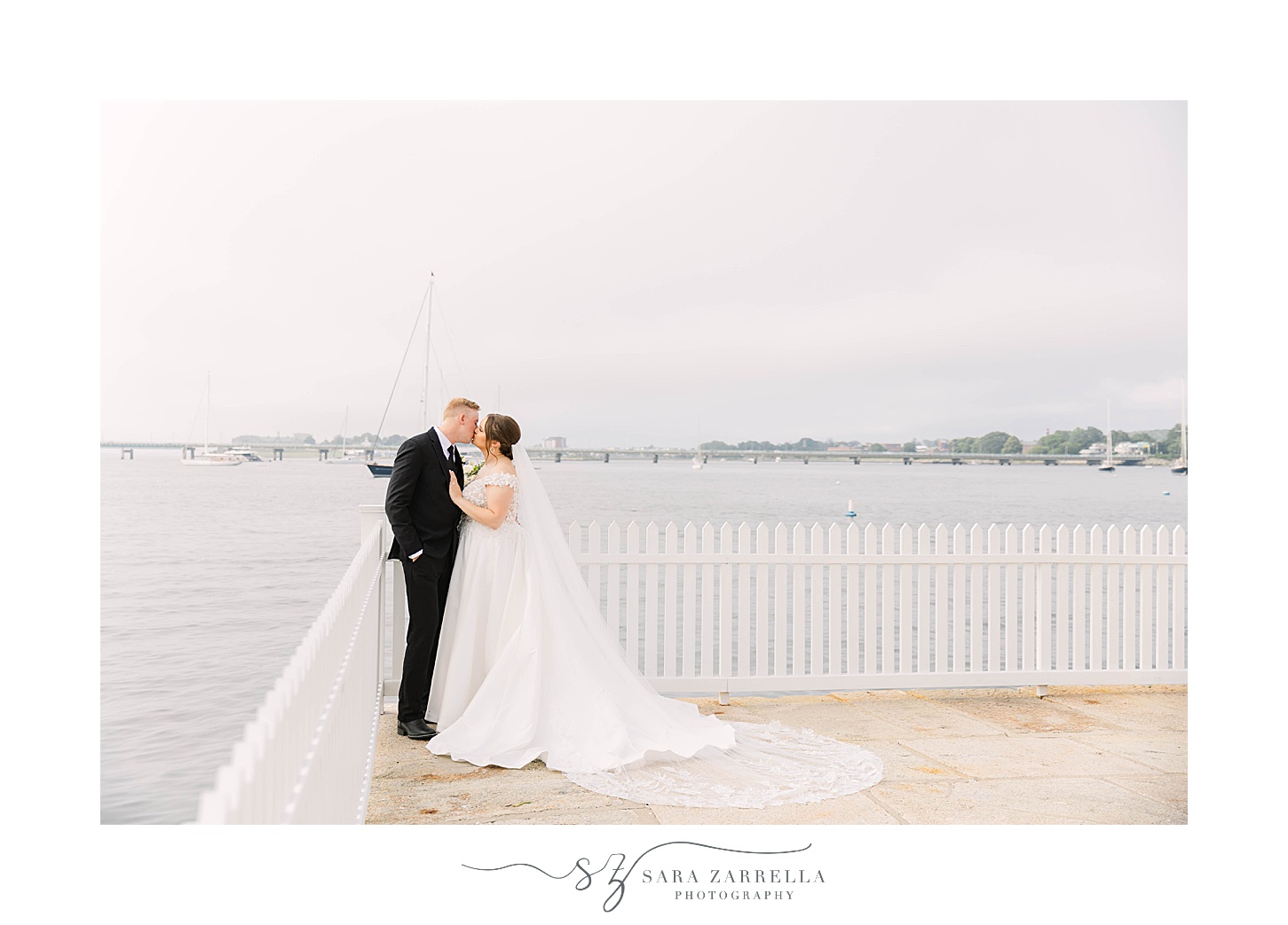 bride and groom kiss in corner of white fence in front of Narragansett Bay