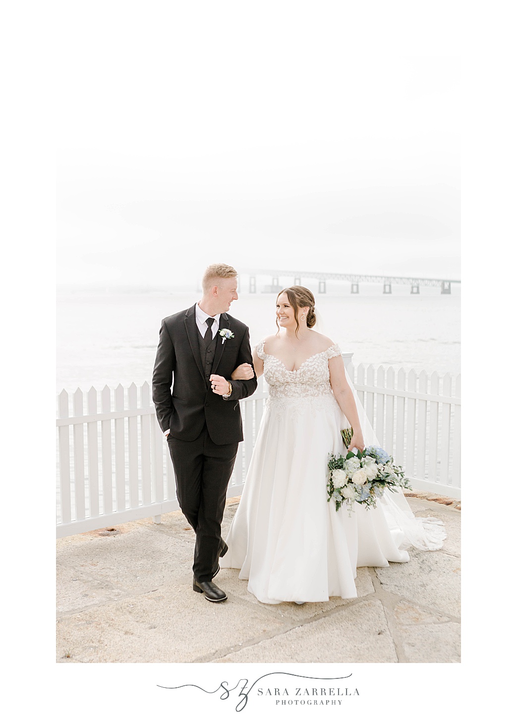 bride and groom hold hands walking away from white fence at the Narragansett Bay