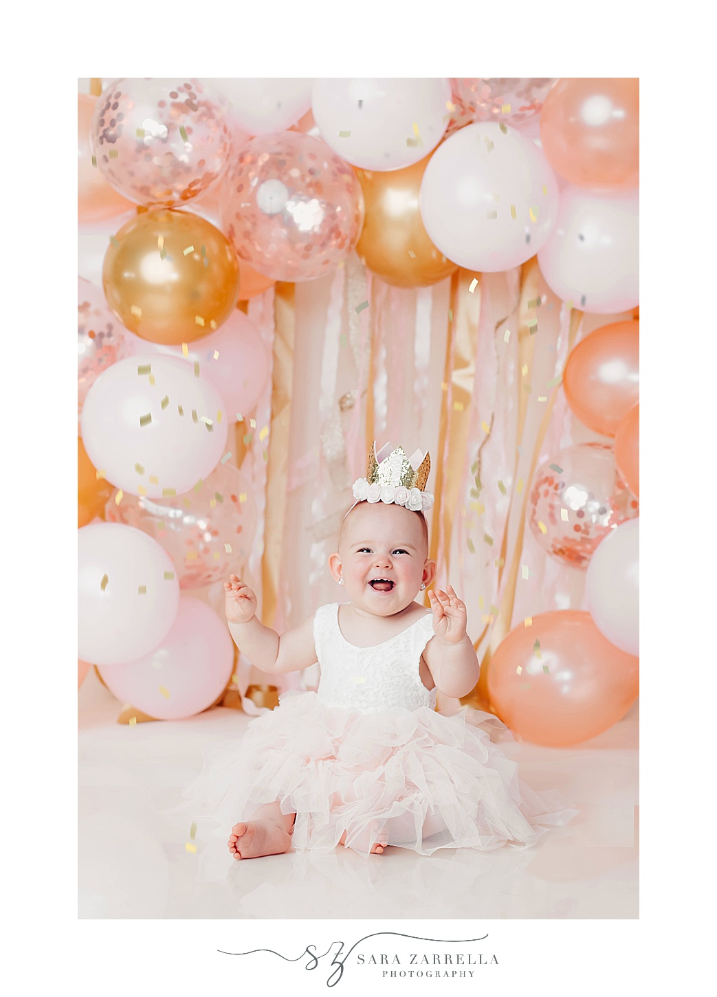 girl laughs in front of pink, orange, and white balloon arch for cake smash portraits with Sara Zarrella Photography