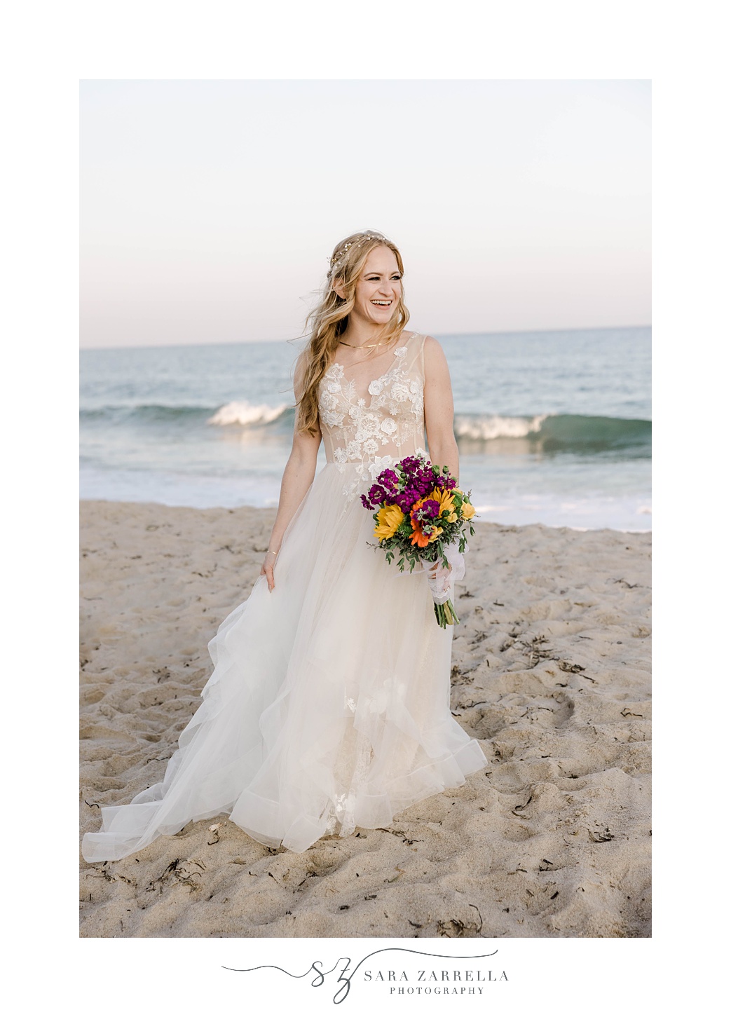 bride stands on beach holding bouquet of bright purple and yellow flowers 