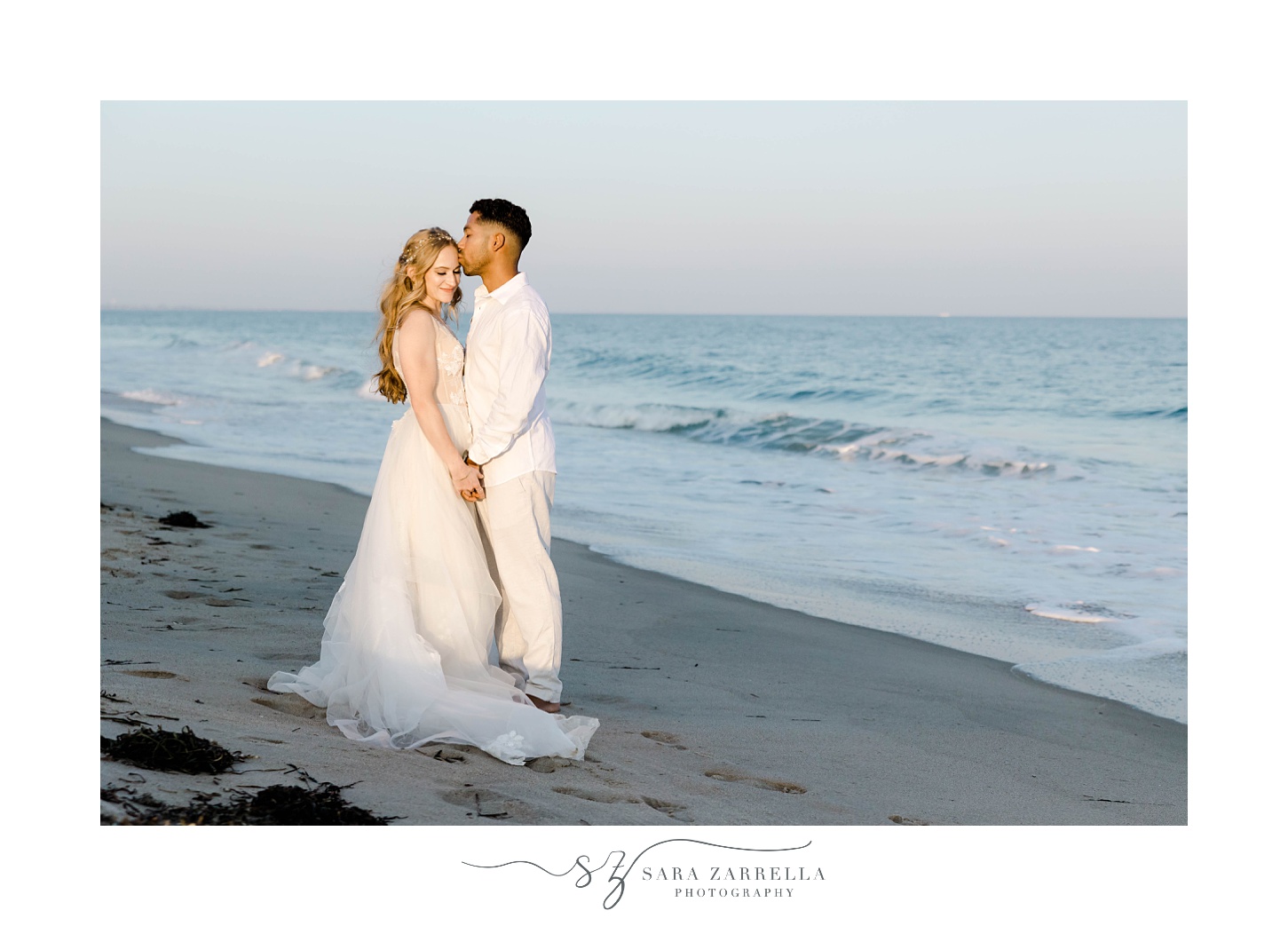 groom kisses bride's head holding hands in front of ocean