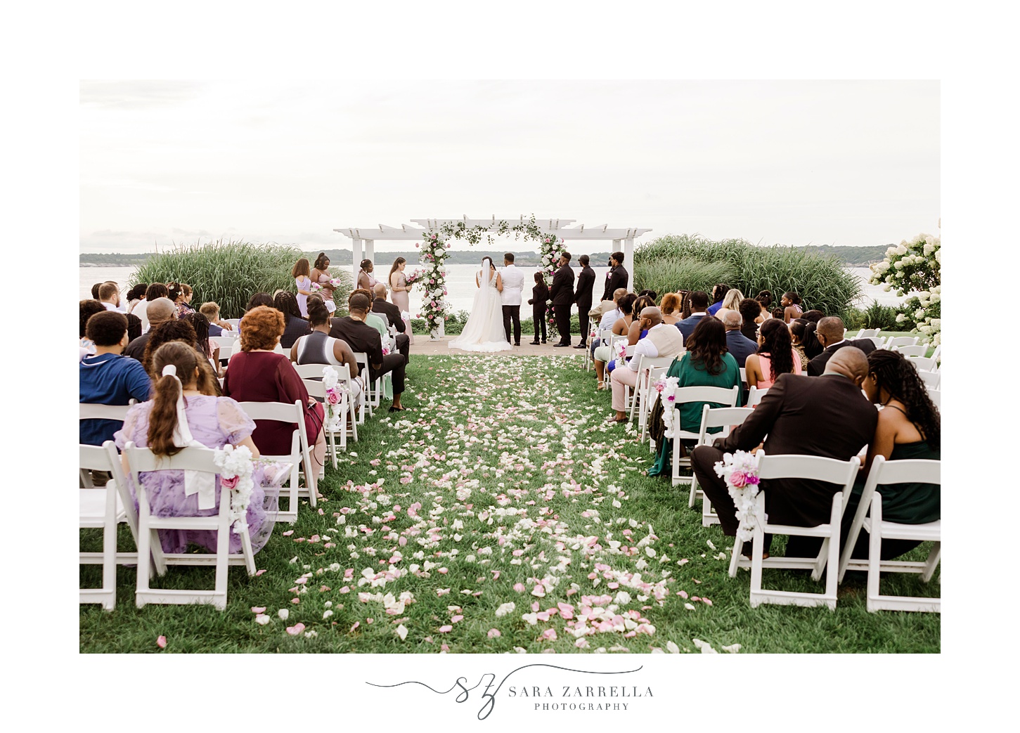 newlyweds stand under white arbor with pink and white flowers at OceanCliff Hotel