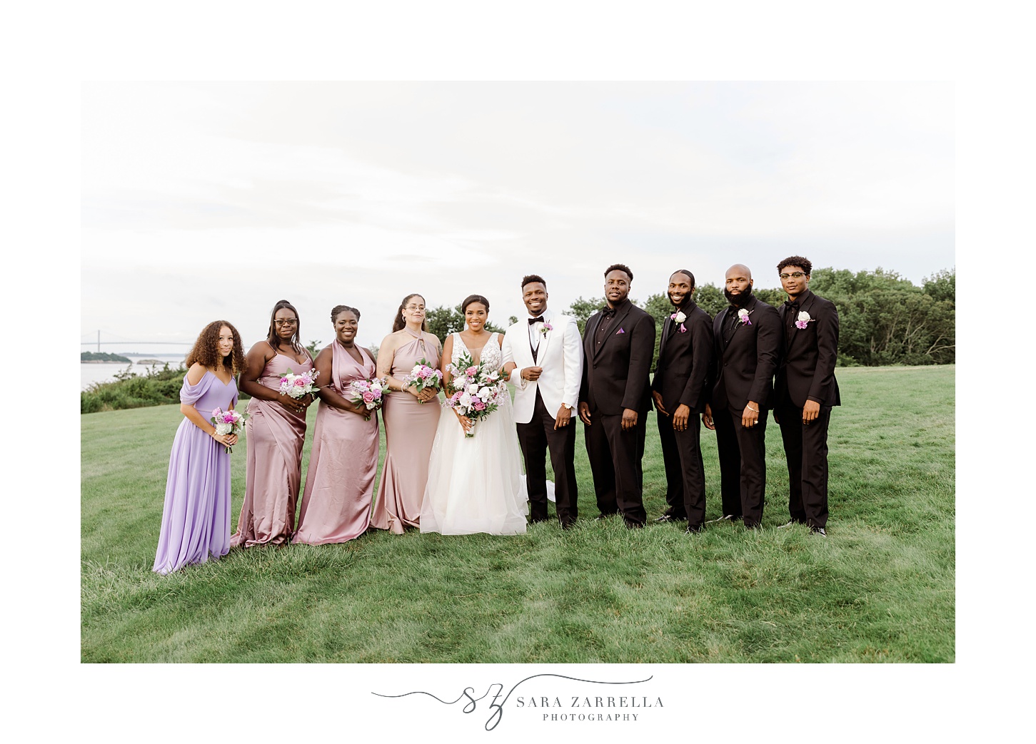bride and groom stand on hill with bridal party in pink and purple dresses and black suits 