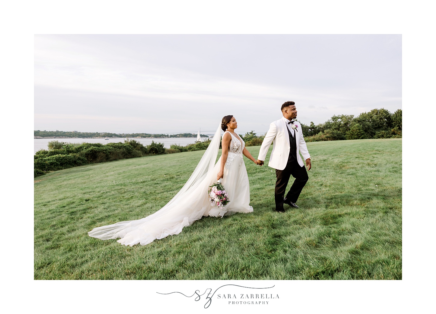 groom in whit suit jacket holds bride's hand walking her on lawn in Newport RI