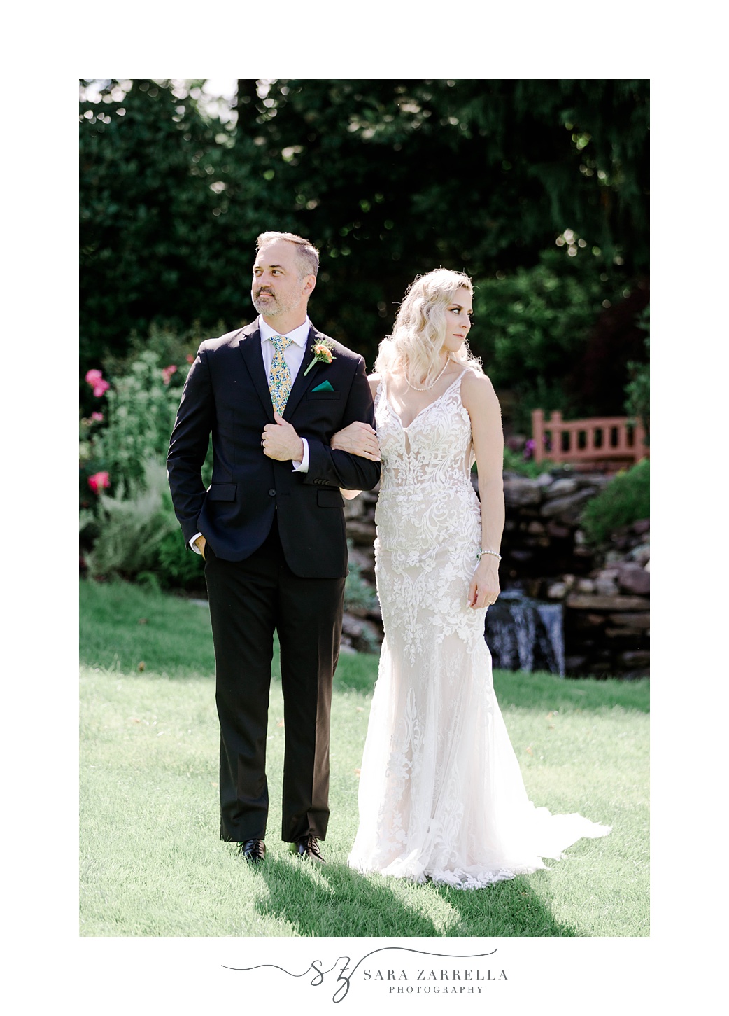 bride and groom stand on lawn linked arms looking over shoulders 