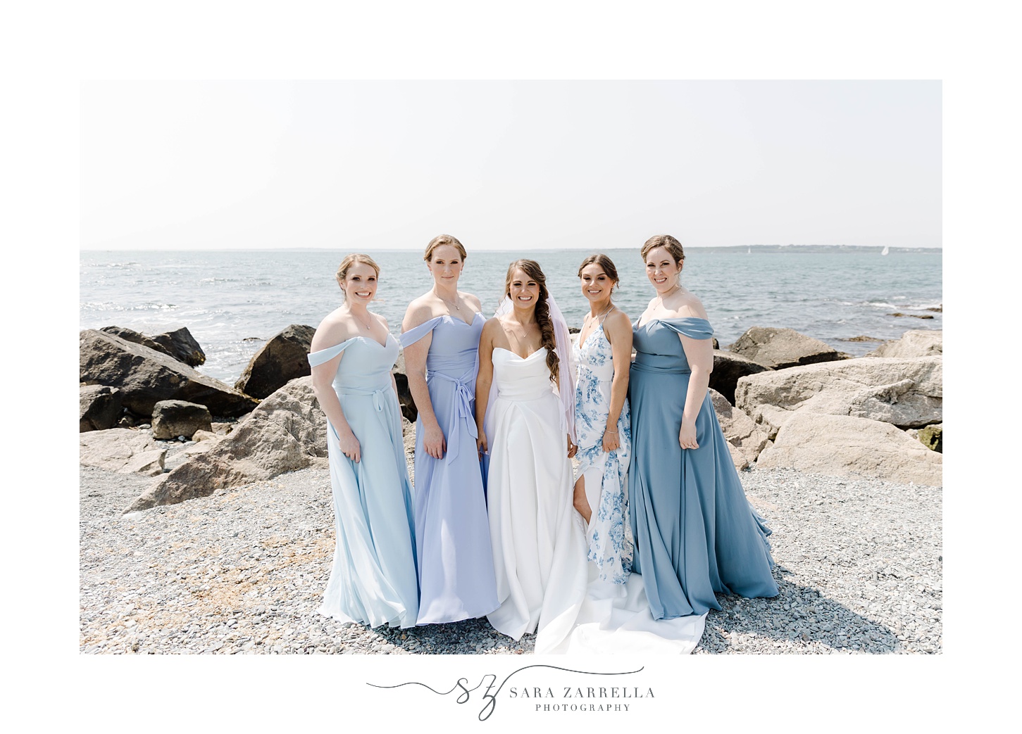 bride and bridesmaids pose in light blue gowns on rocks in Newport RI