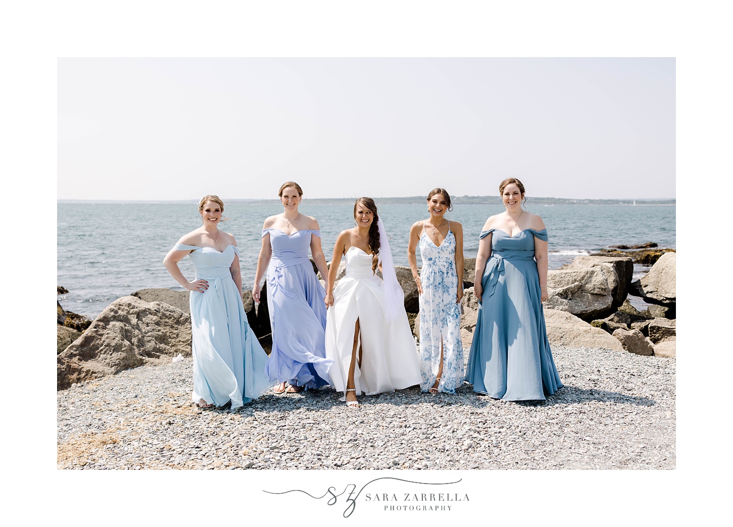 bride and bridesmaids walk along rocks in Newport RI in mismatched blue gowns