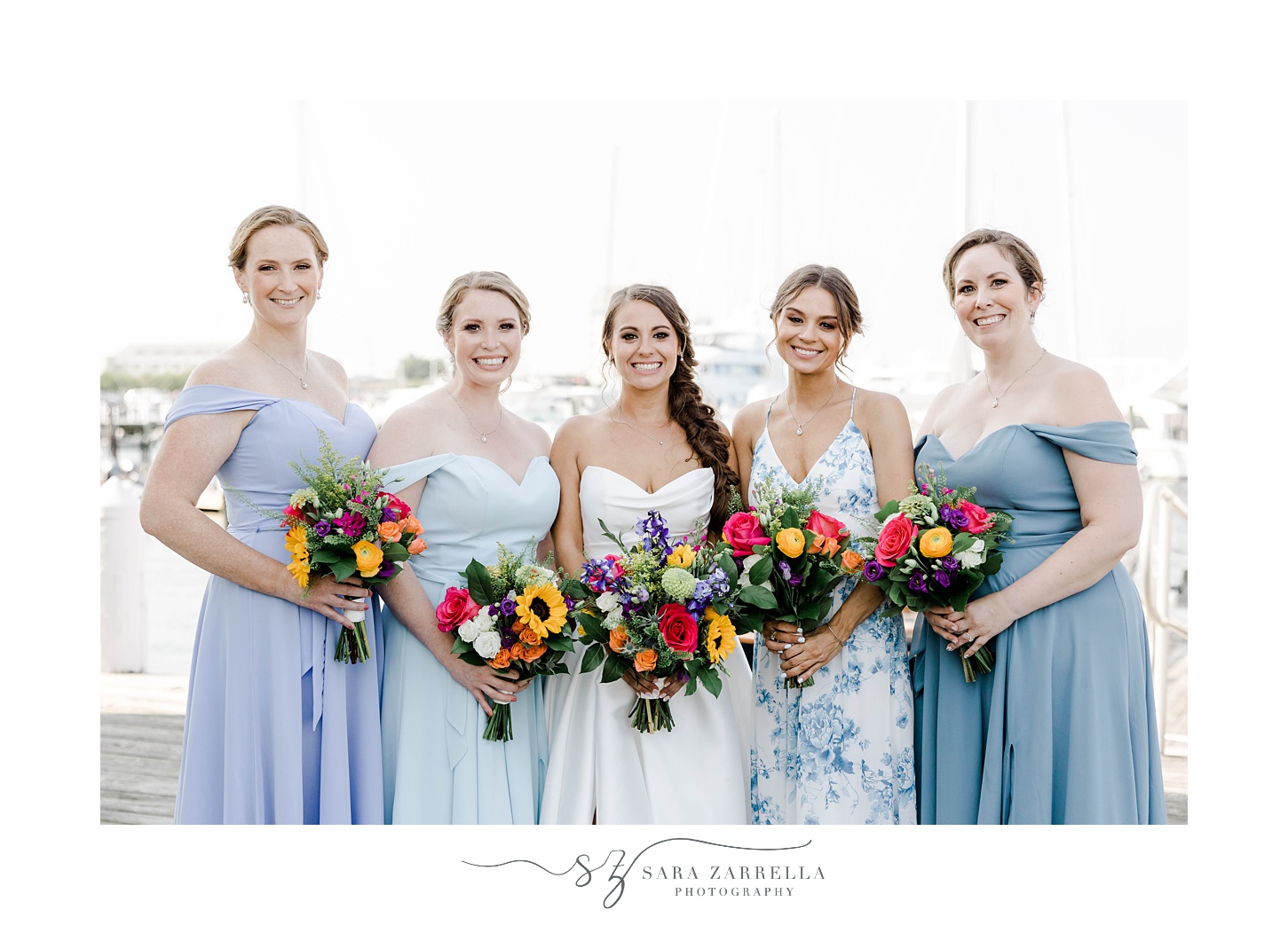 bride stands with bridesmaids in mismatched blue gowns holding bright summer bouquets 
