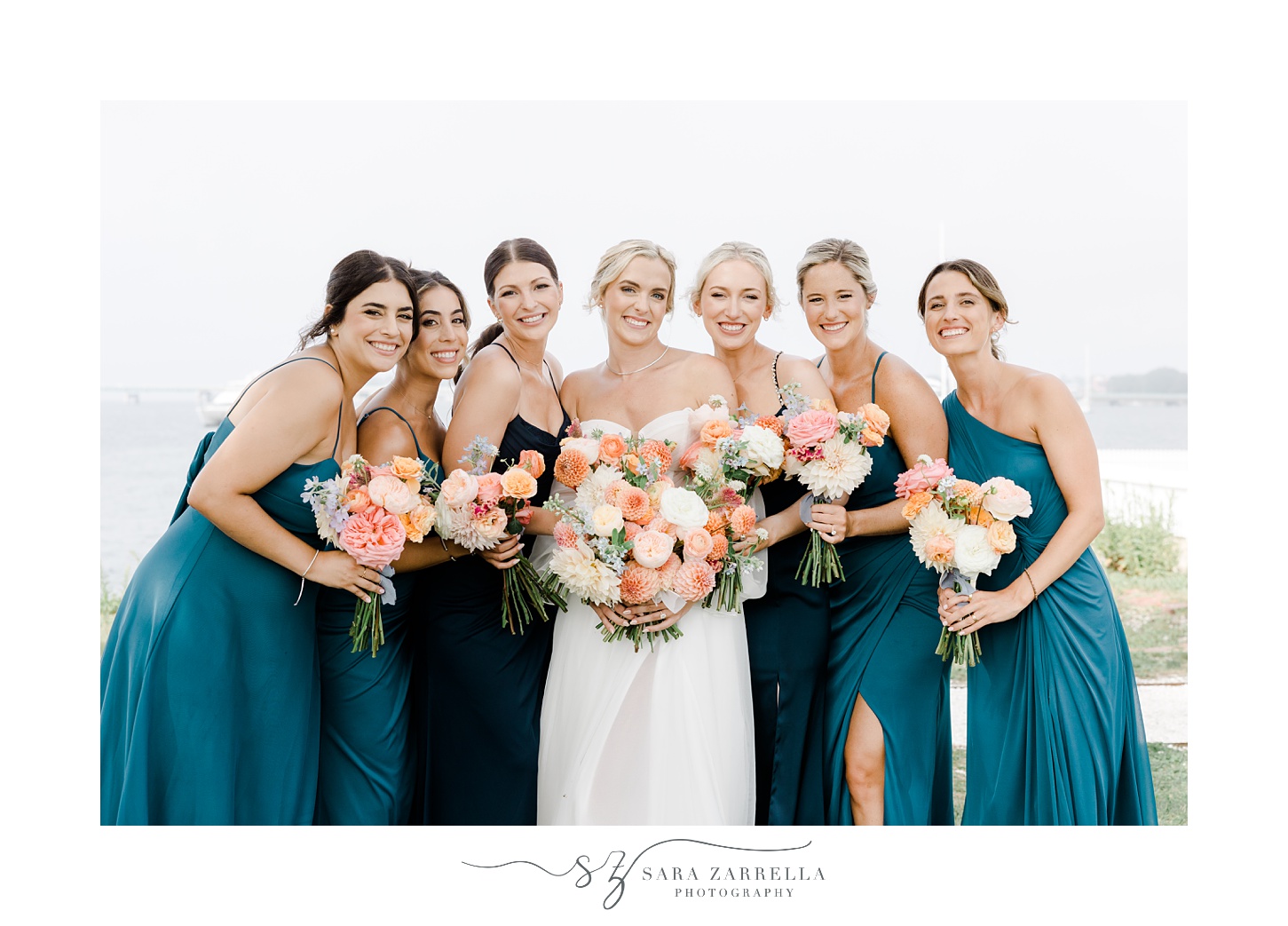 bridesmaids in teal gowns lean into bride in front of Narragansett Bay
