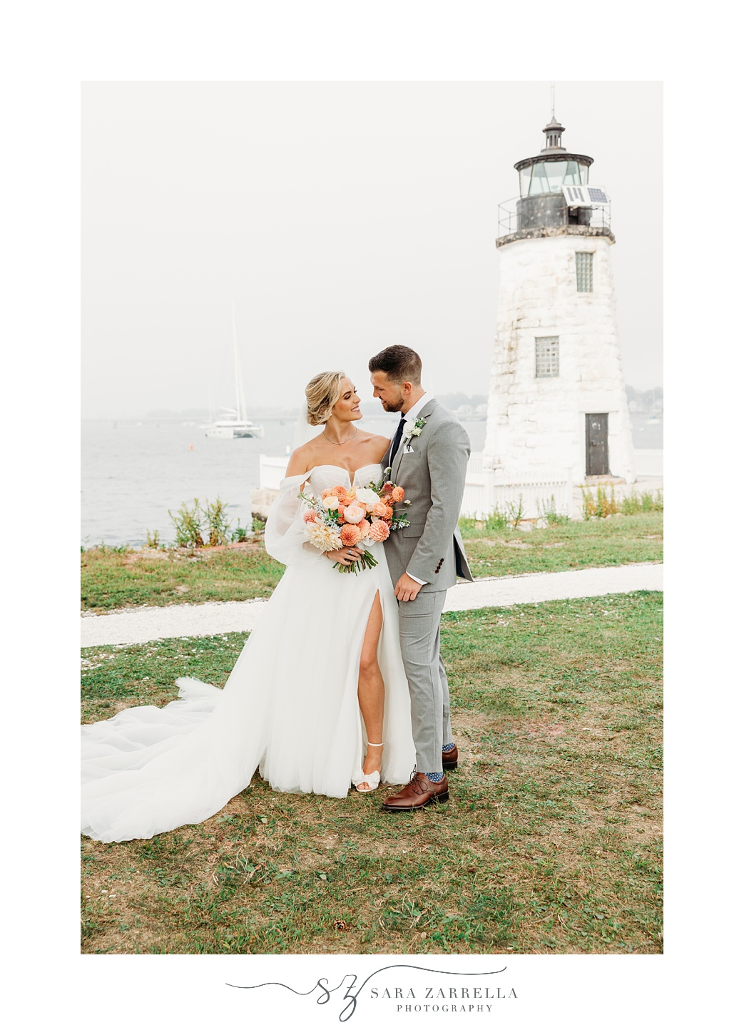 newlyweds stand on lawn in front of Newport Lighthouse holding bouquet of peach and pink flowers 