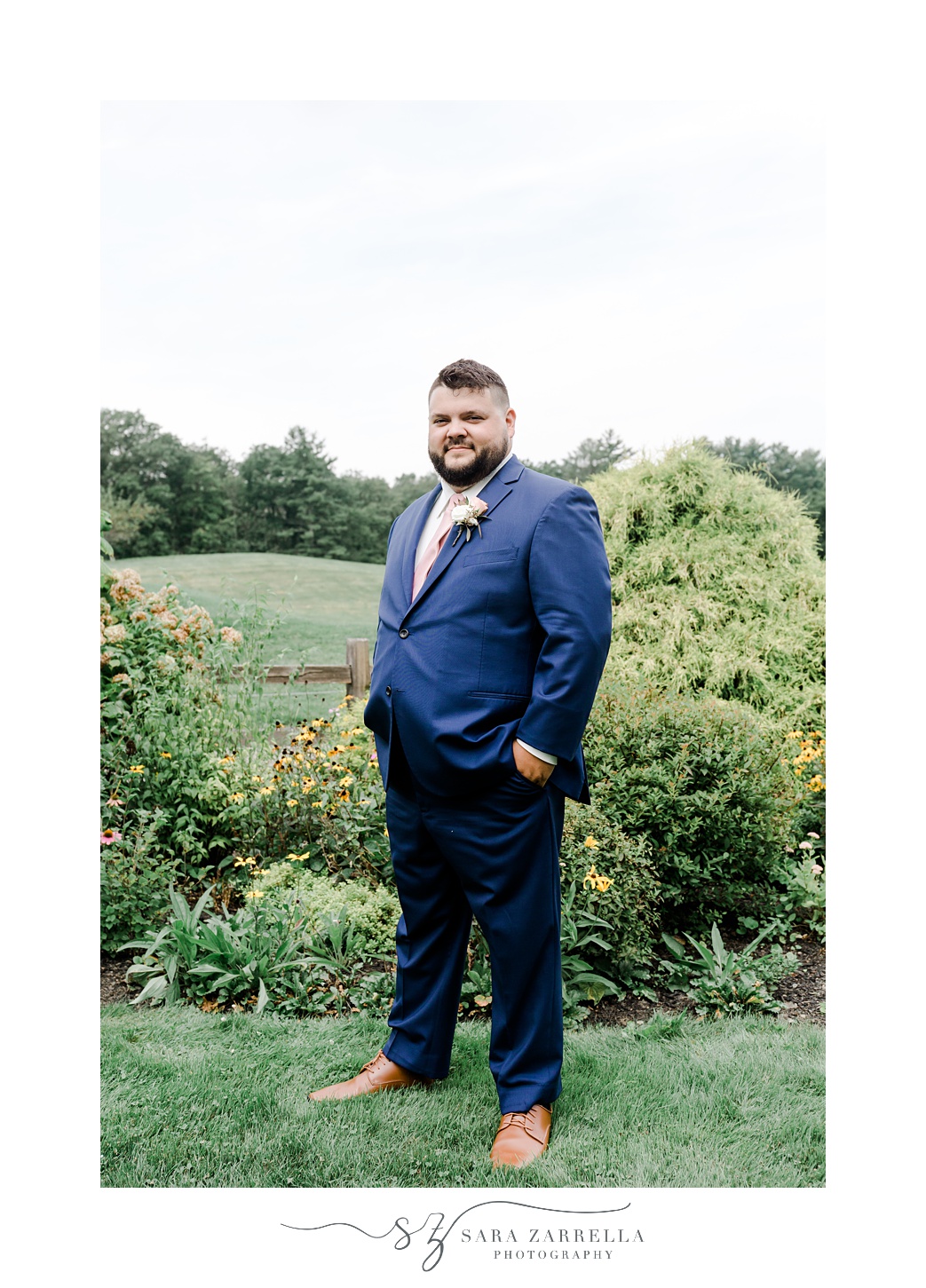 groom stands with hands in pockets of navy suit on Uxbridge MA