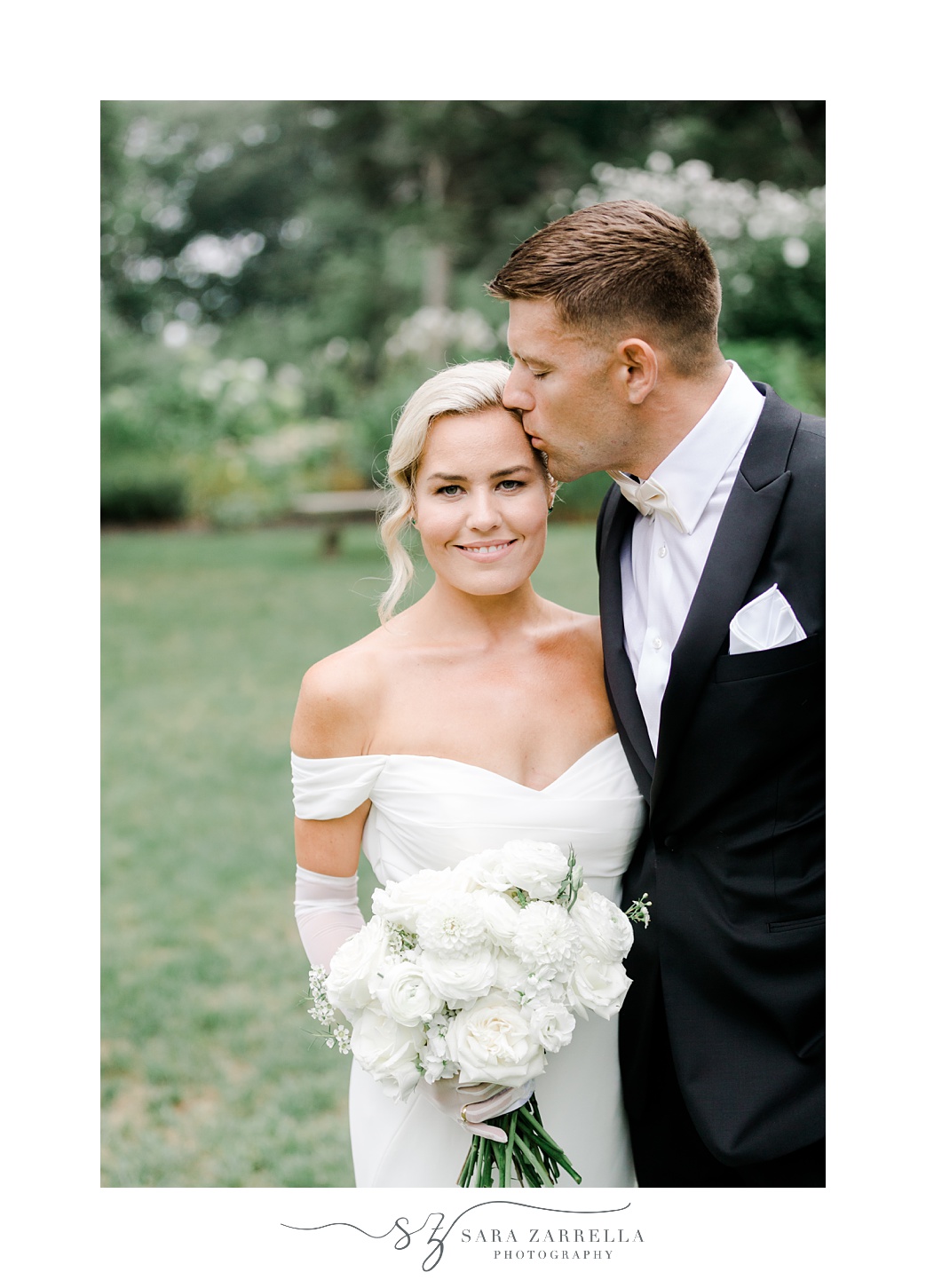 groom leans to kiss bride's forehead in off-the-shoulder gown while holding ivory flowers 