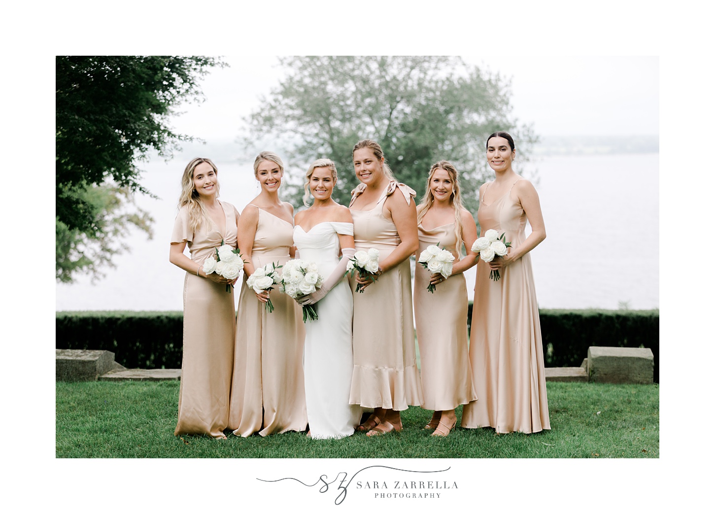 bride poses with five bridesmaids in champagne gowns in front of water at Glen Manor House