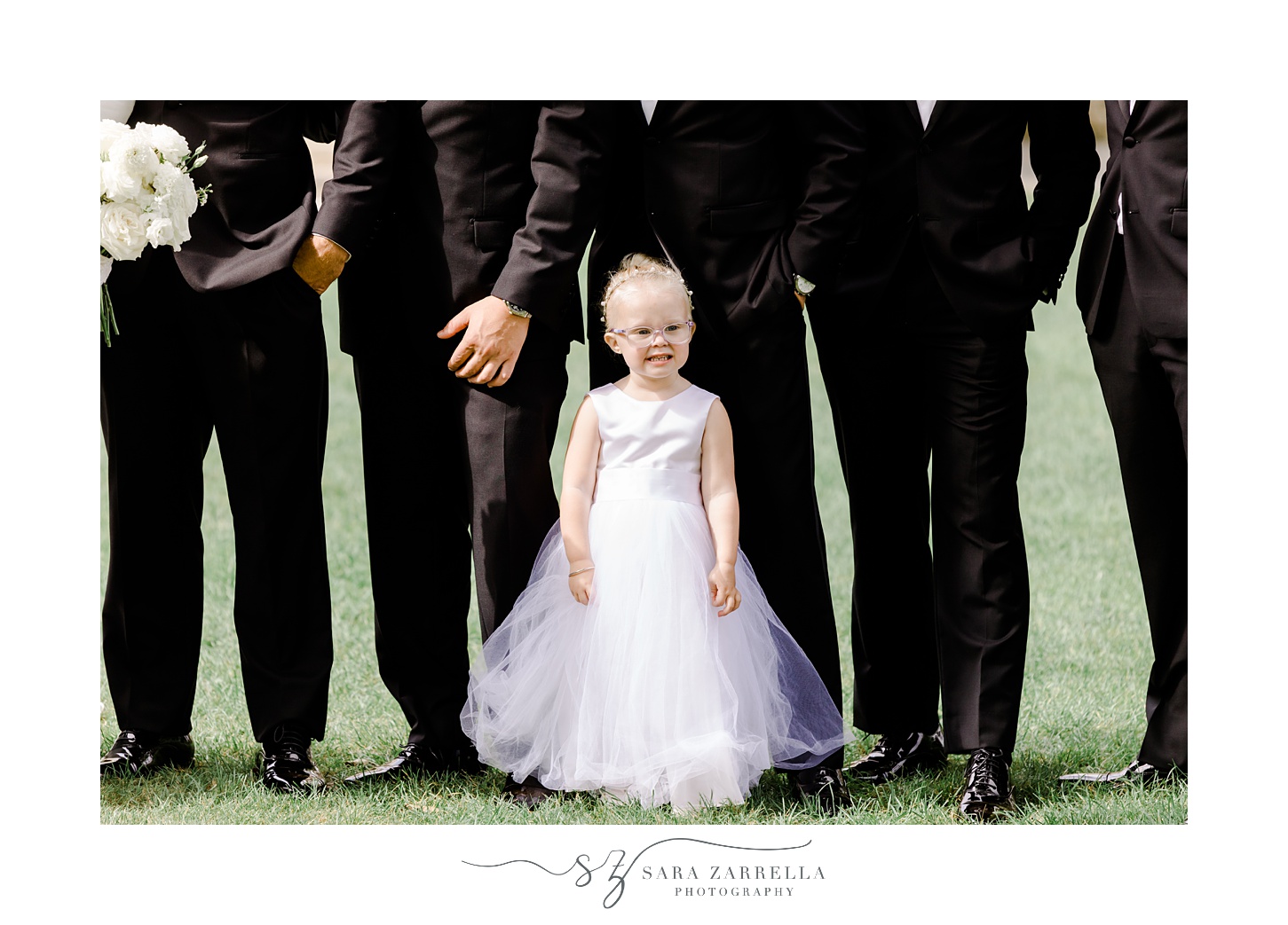 flower girl in white gown smiles in front of groomsmen