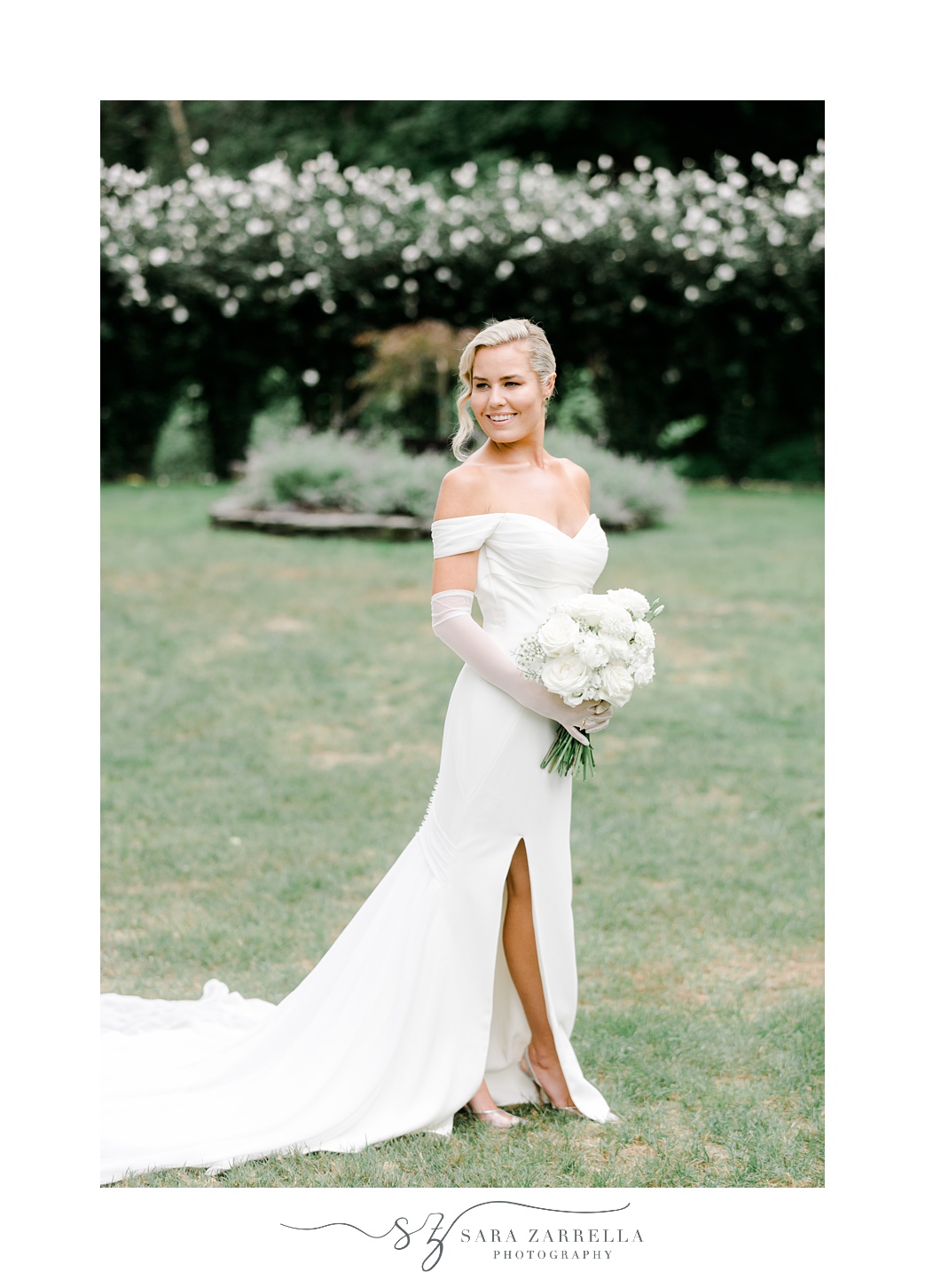 bride stands on lawn holding bouquet of white flowers in white gloves 